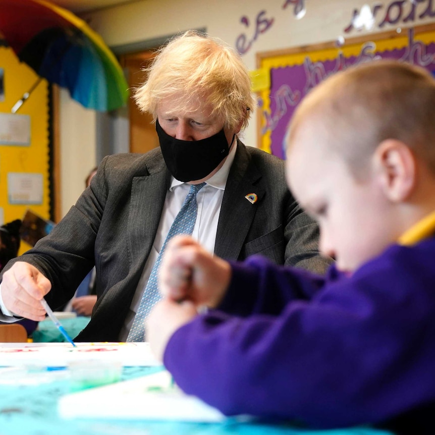 Boris Johnson sits in a chair holding a paint brush as he looks at a piece of paper next to a child who is painting.