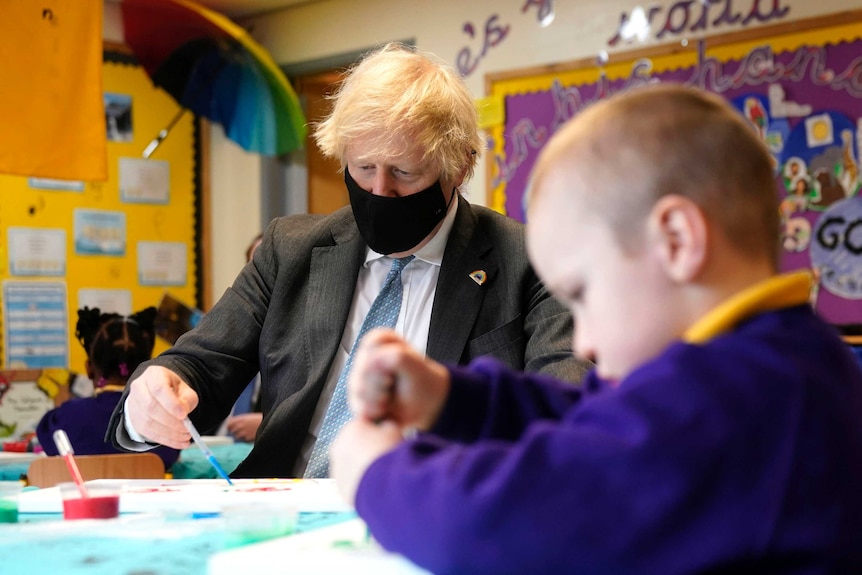 Boris Johnson sits in a chair holding a paint brush as he looks at a piece of paper next to a child who is painting.