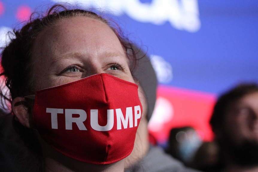 A woman with brown hair wears a red mask with Trump written on it with people behind her.