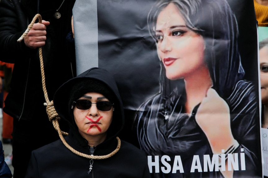 A woman stands with a noose around her neck next to a picture of Mahsa Amini.
