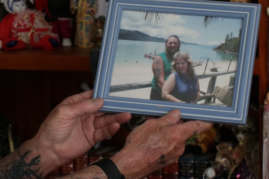 Man's hands holding framed photo of couple on beach, dolls on shelf in background, man has tattoos on arms