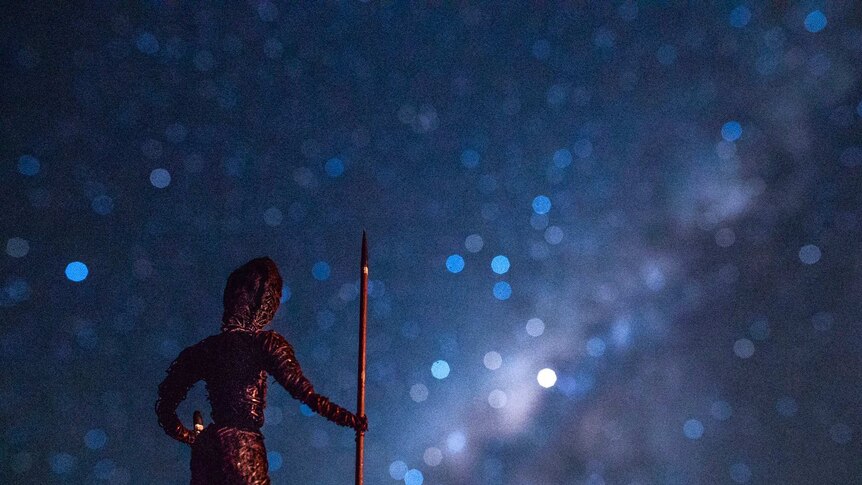 A tiny statue of a man holding a spear looks up to the Southern Cross in a starry night sky.