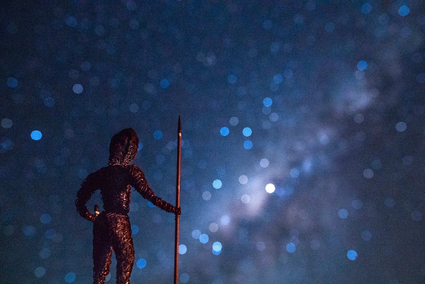 A tiny statue of a man holding a spear looks up to the Southern Cross in a starry night sky.