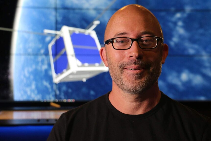 A man with glasses sitting in front of a picture of a satellite in space.