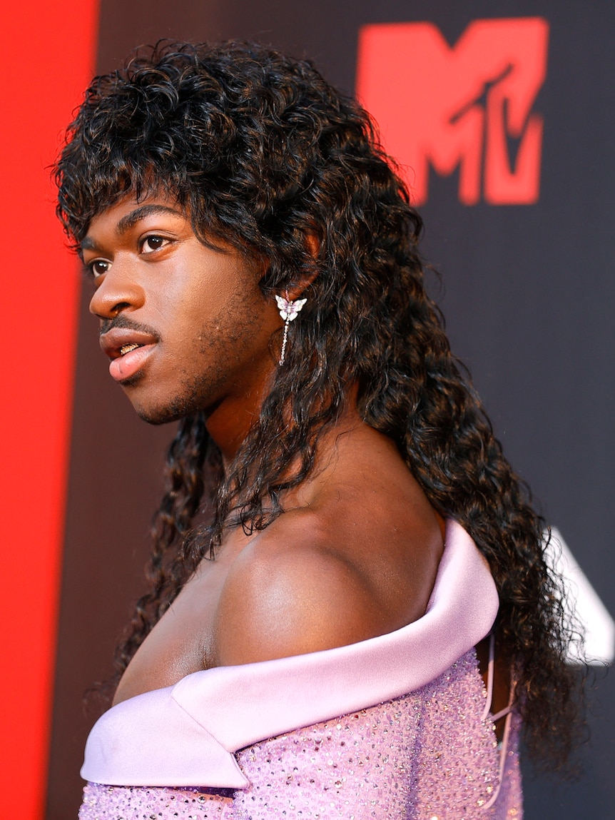 Lil Nas X on the MTV red carpet wearing a purple top and long curly mullet. 