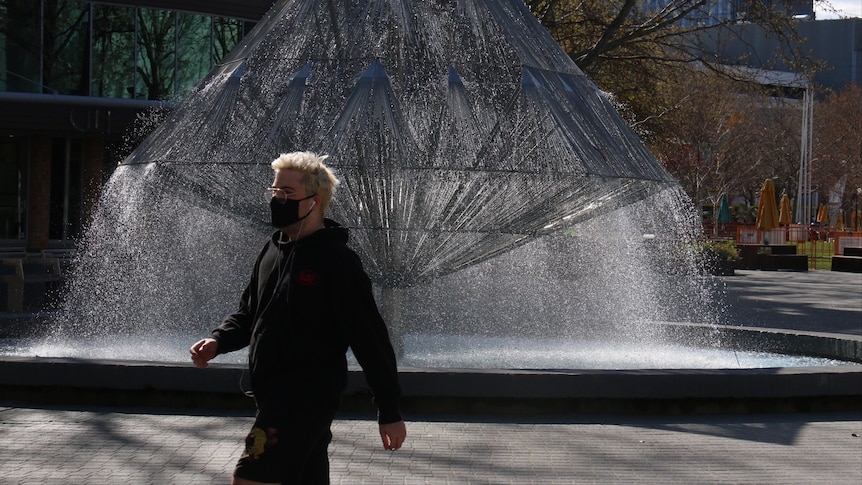 A man in a face mask walks in front of a fountain.