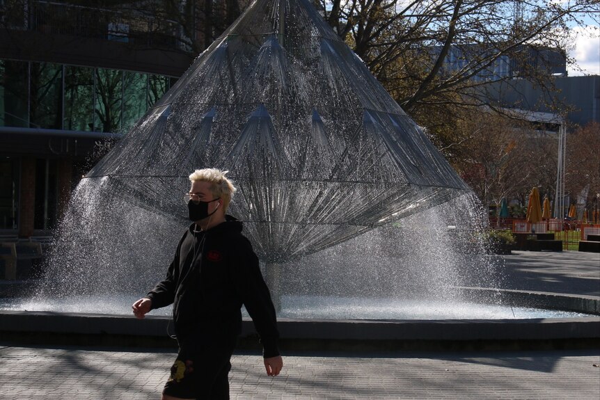 A man in a face mask walks in front of a fountain.