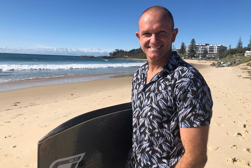 A man in a patterned shirt stands on a beach holding a bodyboard, smiling.