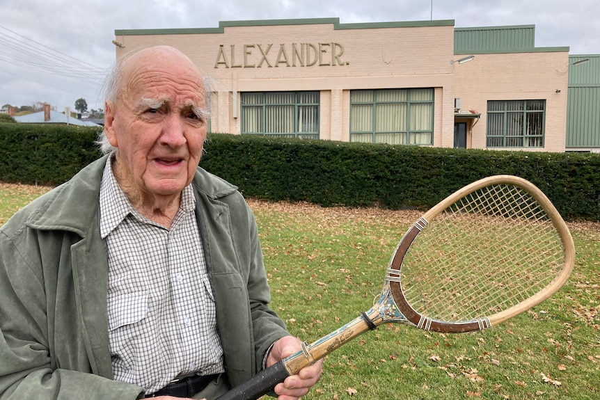 A man stands in front of a building holding a tennis racquet. The building has the word 'Alexander' written on it