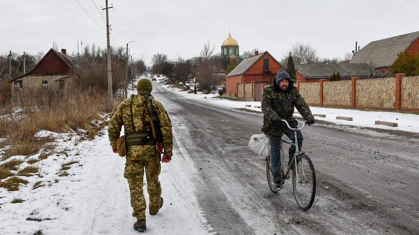 A man riding a bicycle rides past a soldier walking along a snow covered street in a Ukrainian village.