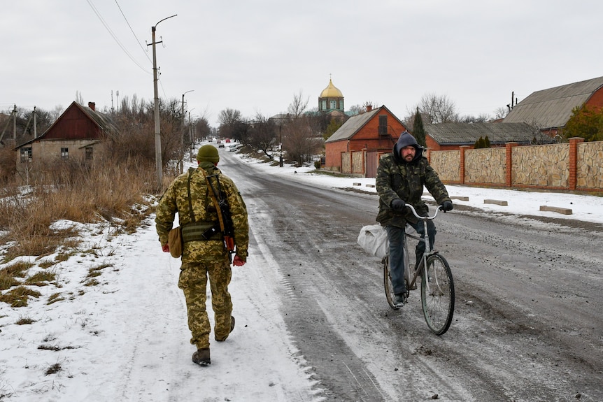 A man riding a bicycle rides past a soldier walking along a snow covered street in a Ukrainian village.