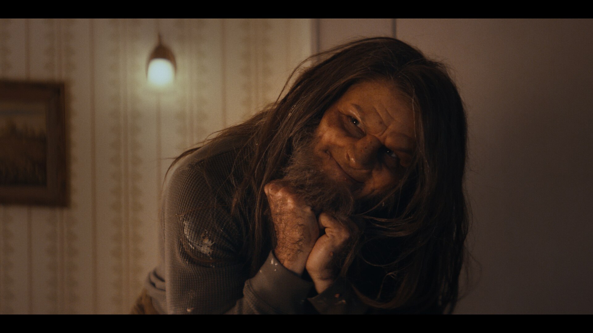 A close-up film still of Tommy Dewey, made up like a monster, smiling, his fists resting under his chin.