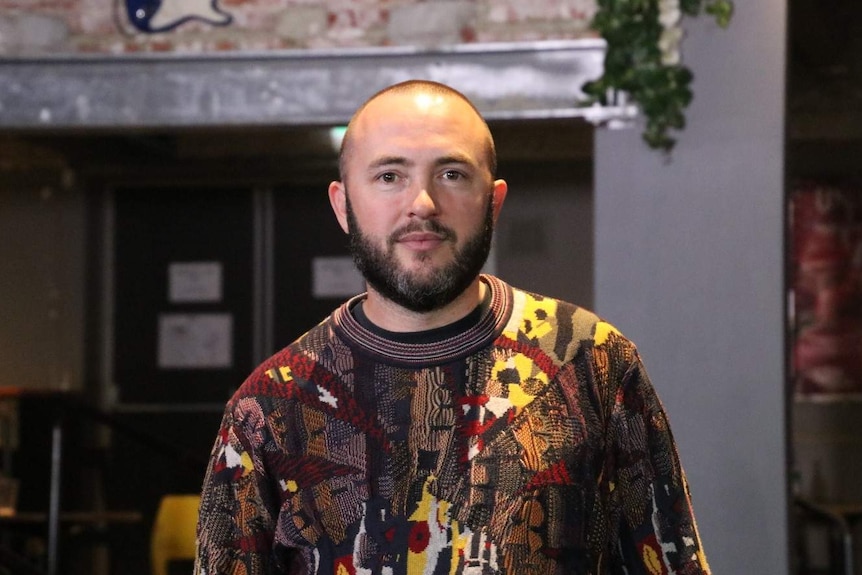 Andrew Ryan wearing a colourful knitted jumper inside a pub with exposed brick and an electric guitar on the wall.