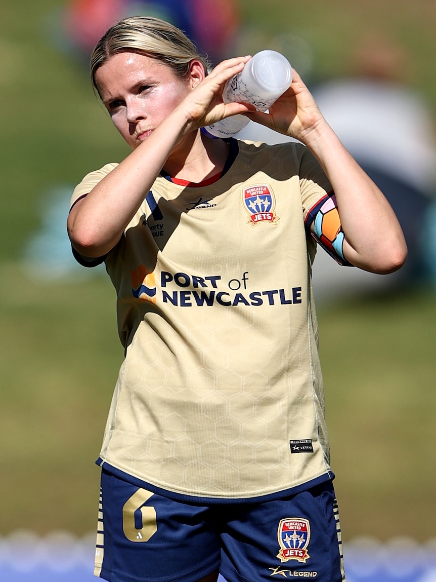 A soccer player wearing a gold shirt squirts a water bottle over her shoulders during a hot game