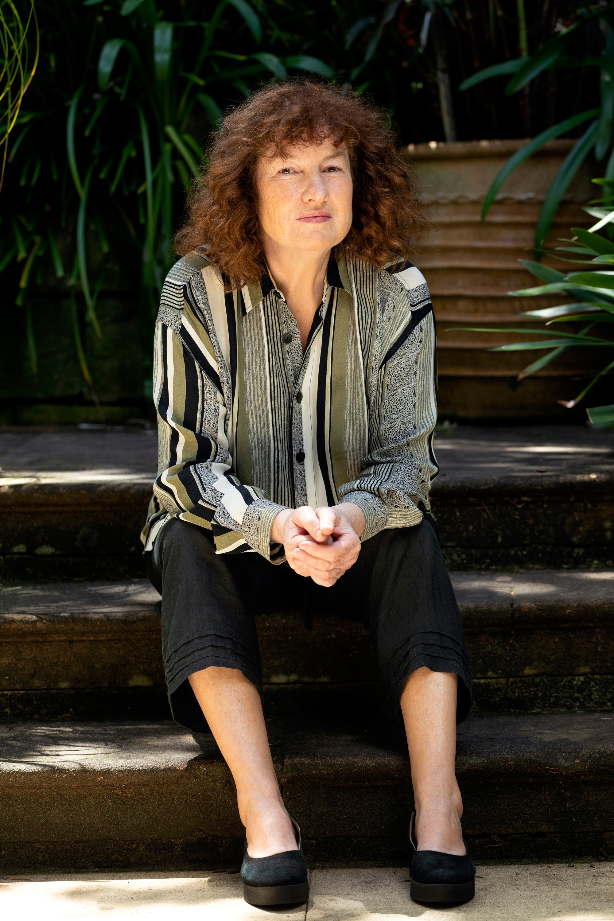 A 60-something woman with brown hair sits on outside steps, in a garden, her hands clasped in her lap