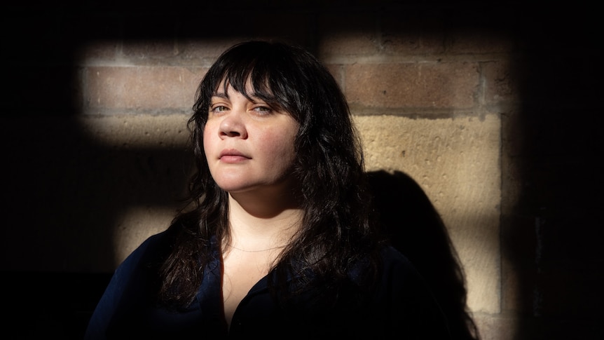 A fair-skinned Aboriginal woman with bangs leans against a brick wall, chin raised, with a defiant expression on her face