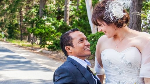 A bride sits on the lap of her groom, who uses a wheelchair. The newlyweds look at each other, smiling.