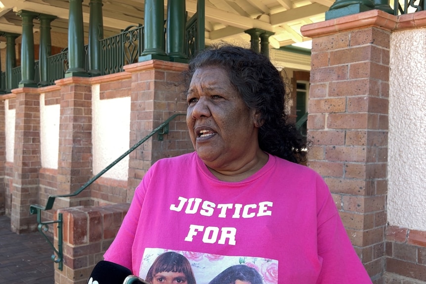 An Indigenous woman in a brightly-coloured T-shirt stands speaking in front of a court building.