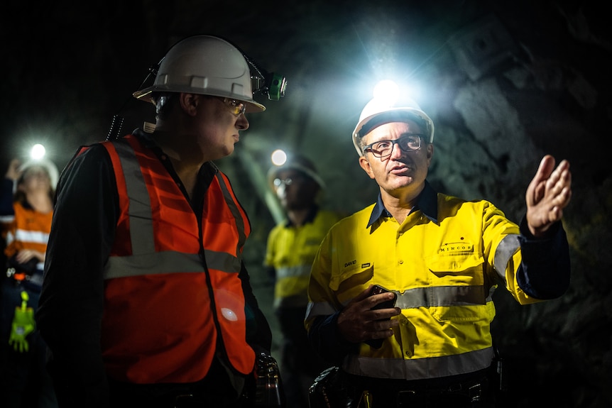 A group of men explore an underground wearing bright coloured shirts and hard hats with lights on them.