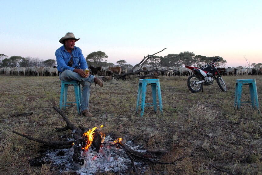 A man sits on a blue stool beside a motorbike, in front of a small campfire
