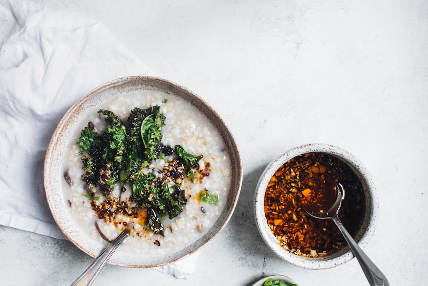 A bowl of congee from our easy recipe with kale and chilli topping sits beside a small bowl of chilli oil.