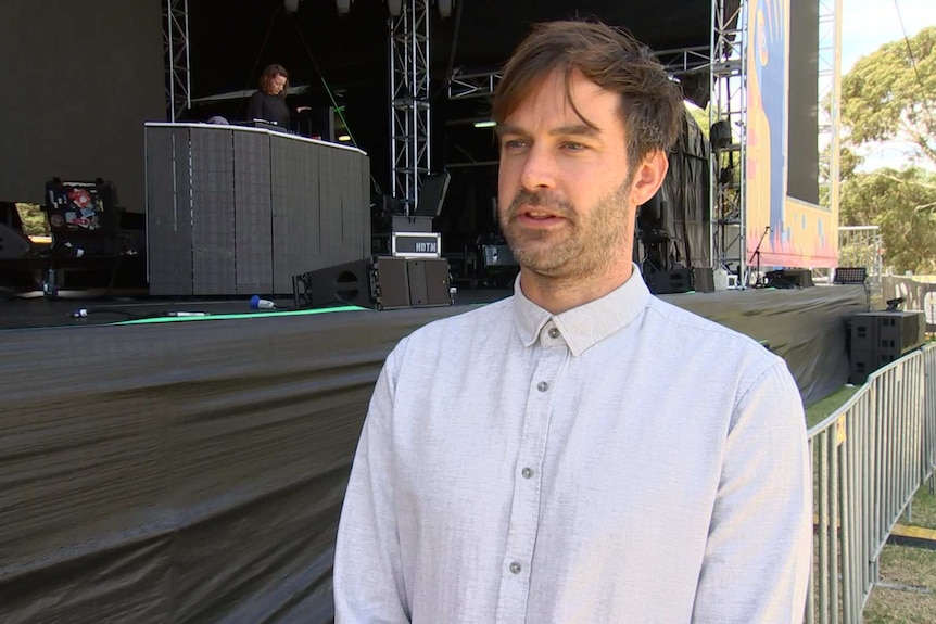 A man in a grey collared shirt, standing in front of a stage, where people are setting up for a band.