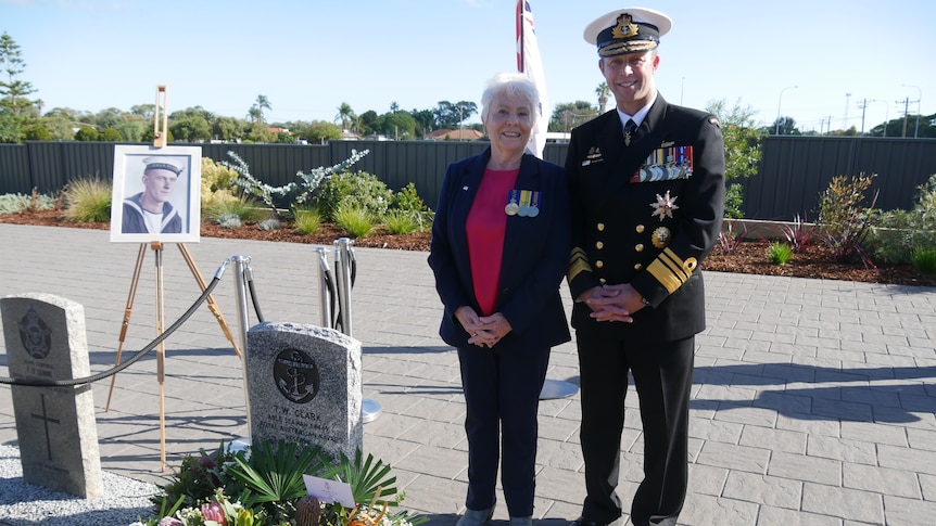 A woman wearing medals stands alongside a man in Navy uniform near a headstone.