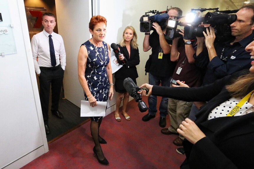 Pauline Hanson speaks to several reporters wile wearing a blue and white dress with pearls. James Asby stands behind her.