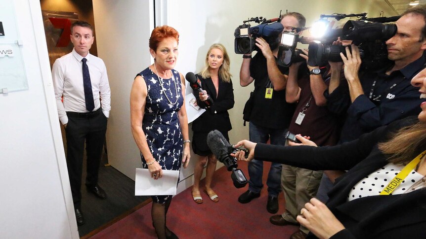 Pauline Hanson speaks to several reporters wile wearing a blue and white dress with pearls. James Asby stands behind her.