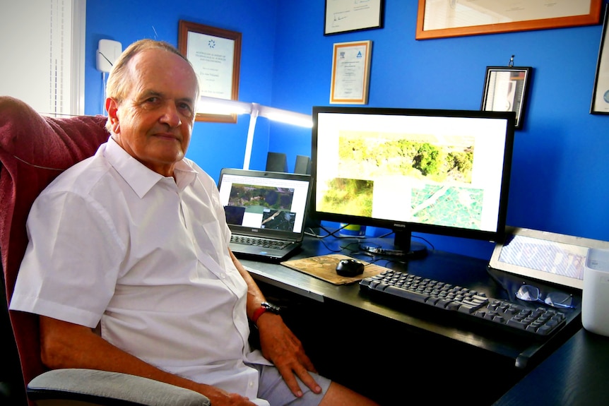 A scientist sits at a desk with a computer of data behind him. 