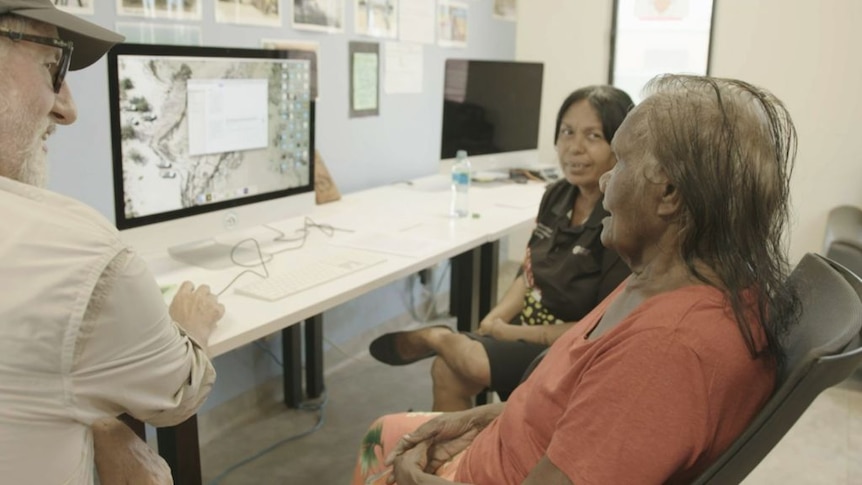 A woman sits in front of a computer screen while a man next to her holds the mouse. A woman is on her right looking at her.