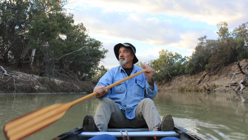 A man with a beard and a fishing hat paddles a canoe on a brown river.