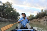 A man with a beard and a fishing hat paddles a canoe on a brown river.
