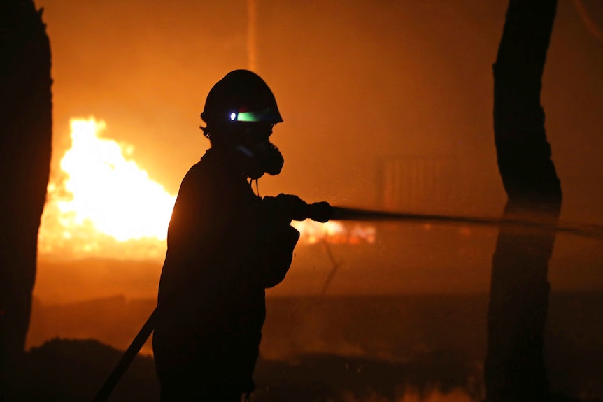 A silhouette of a firefighter wearing a mask and spraying water from a hose against a background of orange flames