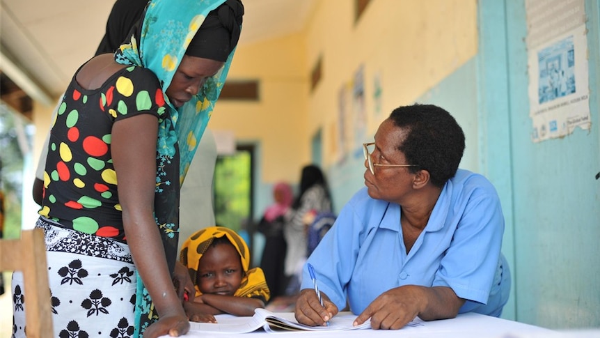 A woman speaks to a man sitting at a desk filling out a form as a child stands at her side.