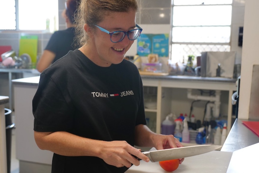 Emily Slotosch in kitchen wearing a black t-shirt, blue and purple glasses, holding a large kitchen knife and slicing a tomato.