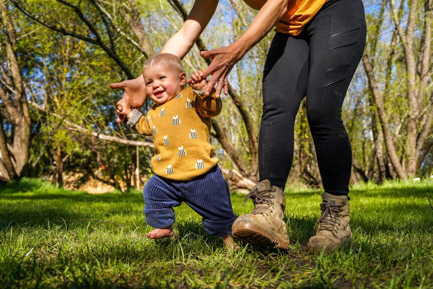 A baby girl supported by her mother's hands runs across a grassy field laughing.