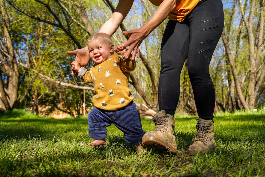 A baby girl supported by her mother's hands runs across a grassy field laughing.