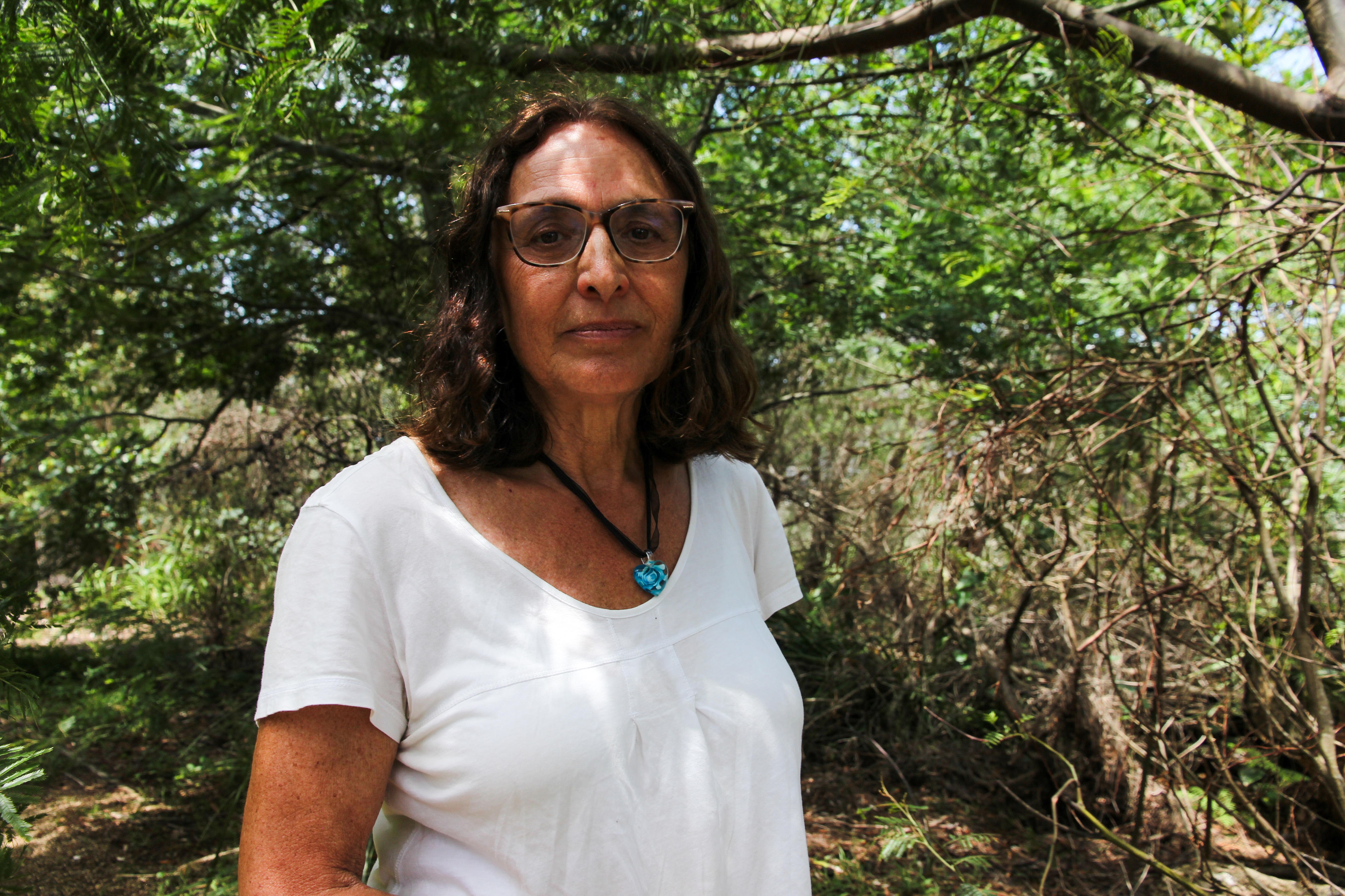 A middle-aged woman wearing glasses and a white t-shirt stands in the middle of a bush.