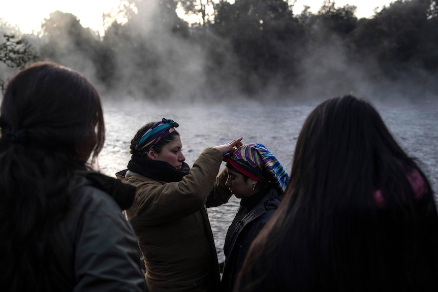 A woman holds her hands over another woman's head in front of a misty river as two more people watch on 