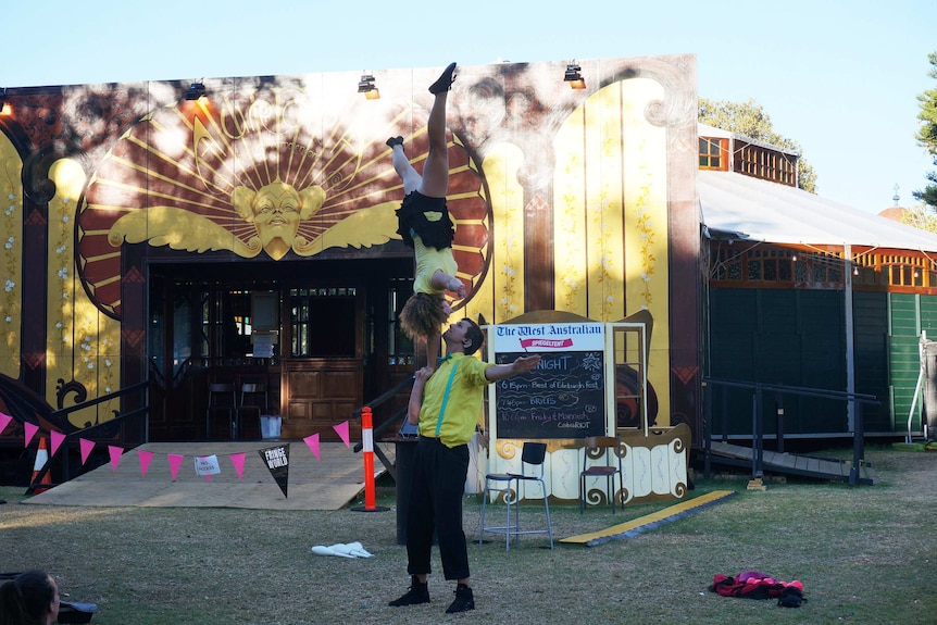 A Fringe street performer balances while doing a handstand on a man's hand.