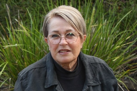 A lady with short blond hair and round wirerimmed glasses in a dark shirt and jacket in front of a garden