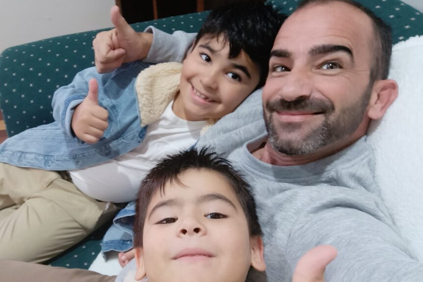 Steven Buhagiar and his sons sitting on a couch and giving the thumbs up while smiling.