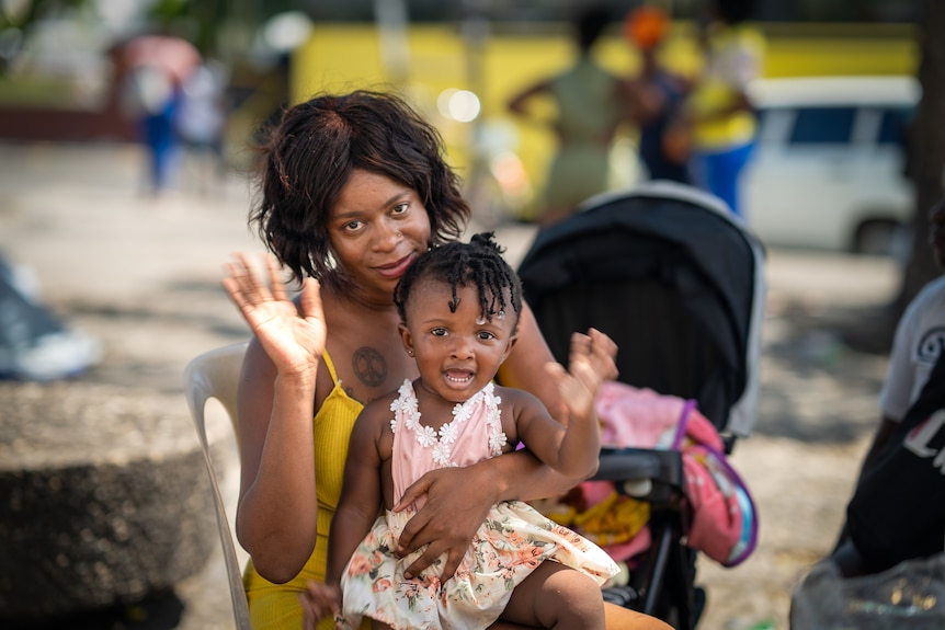 A woman holds a little girl on her lap, as both wave for the camera.