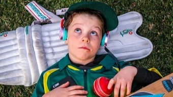 A young boy lies on the grass with earphones in, surrounded by cricket gear.