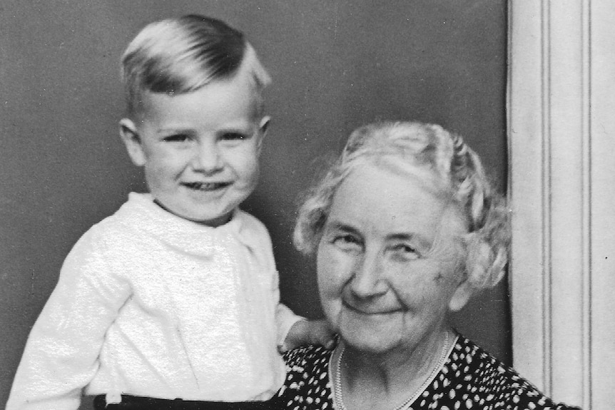 A black and white photo of a young boy standing with his grandmother. 
