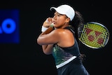 A female tennis player in a dark top plays a two-handed shot during a night match.