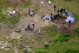 An aerial view of a field there a group of investigators look at an excavated hole near a fire.