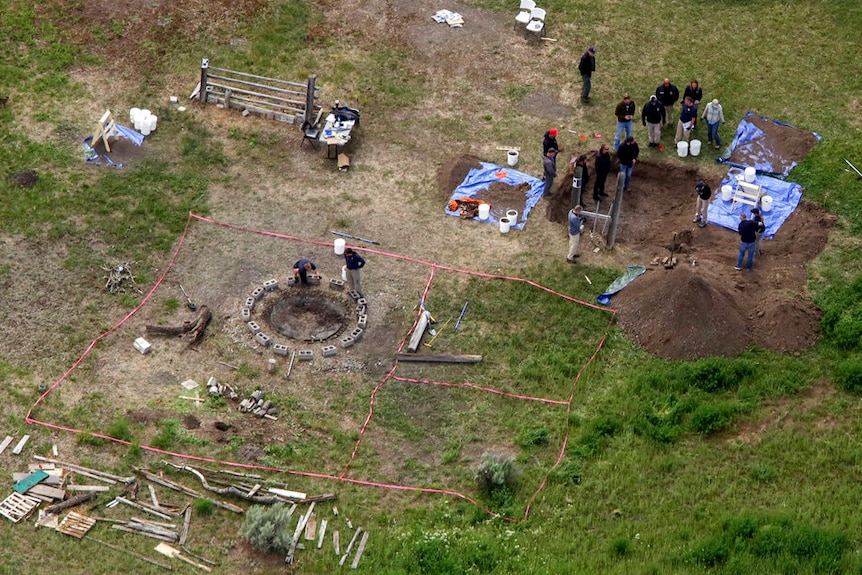 An aerial view of a field there a group of investigators look at an excavated hole near a fire.