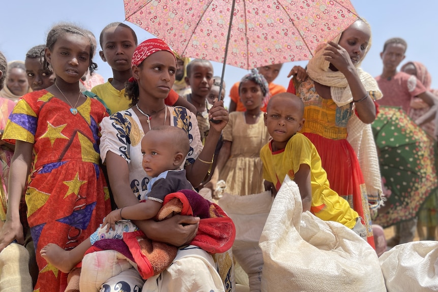 Children gather around woman holding an umbrella for shade while holding a baby on her lap.